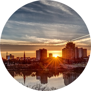 The sun setting over a city with high-rise buildings, and a river in front.