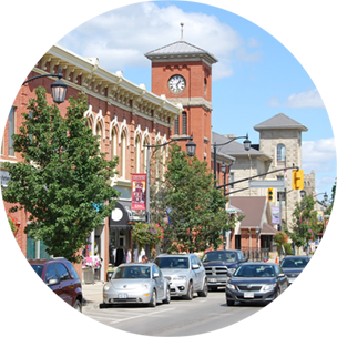 Main street with lots of cars, running parallel to a terracotta coloured building with a clock tower in Milton, Canada.