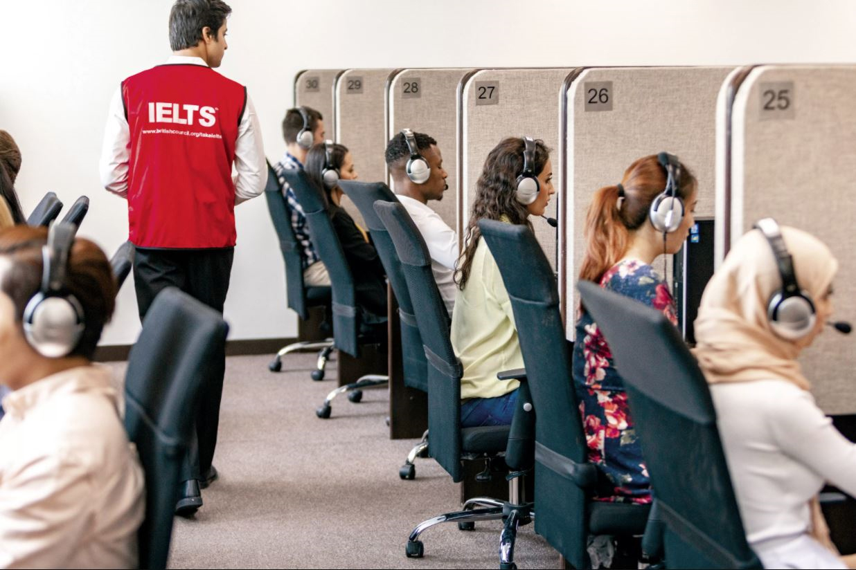 A group of test takers sat in booths with headphones on, sitting IELTS on computers.