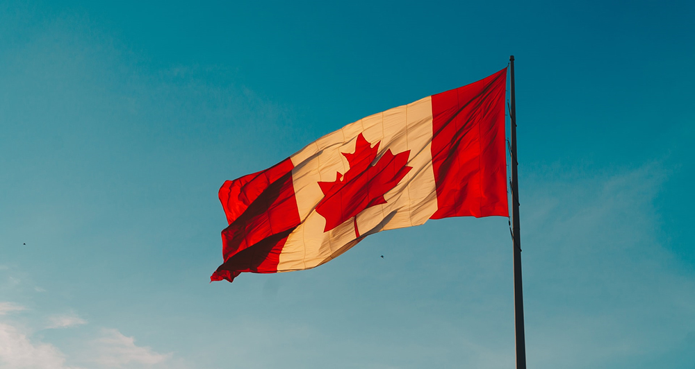 Canadian flag blowing in the breeze on a flagpole, in front of a blue sky.