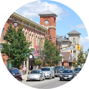 Main street with lots of cars, running parallel to a terracotta coloured building with a clock tower in Milton, Canada.